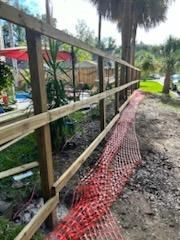 A wooden post-and-rail fence stands beside a fallen orange construction safety fence on a dirt path near trees.