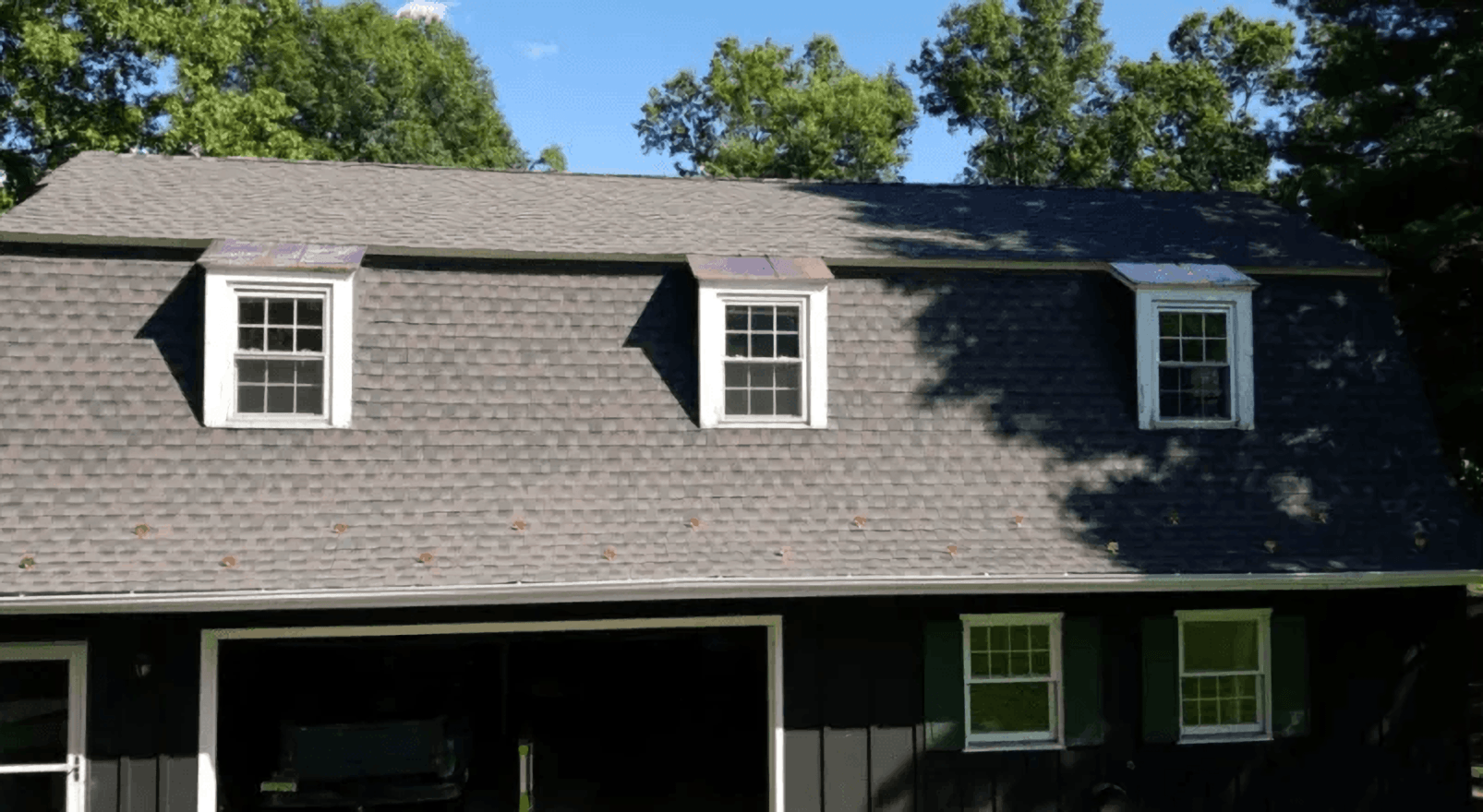 Two dormer windows on a dark shingled house under a clear blue sky, with trees behind it