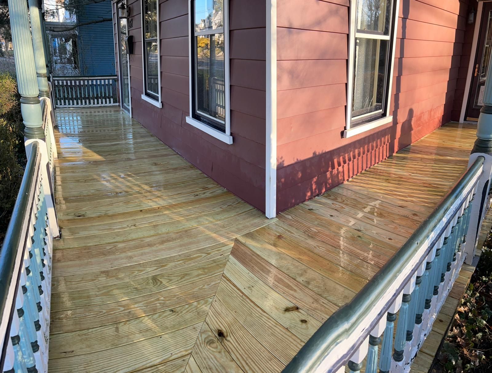 Wooden deck walkway beside a red house, with a metal railing and steps leading to a porch.