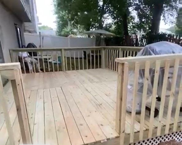 Wooden backyard deck with railing, trees, and a covered grill or furniture piece on the right.