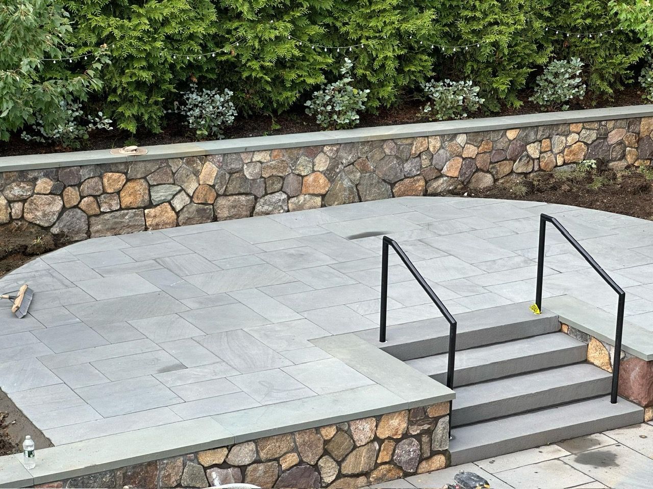 Stone terrace with steps and black handrails beside a low stone wall and greenery