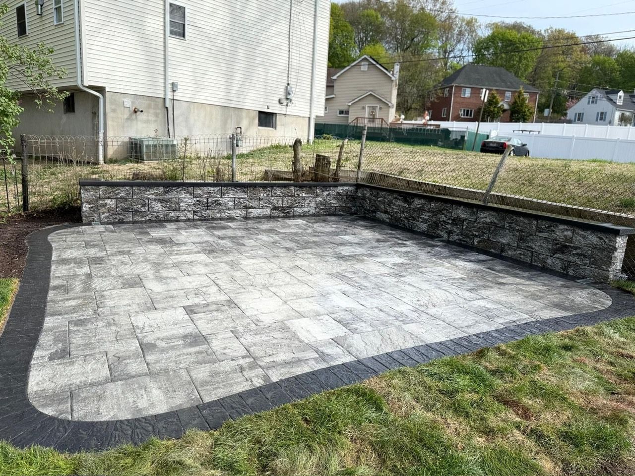 Backyard paved patio with gray stone pavers, black edging, and grass border beside a house
