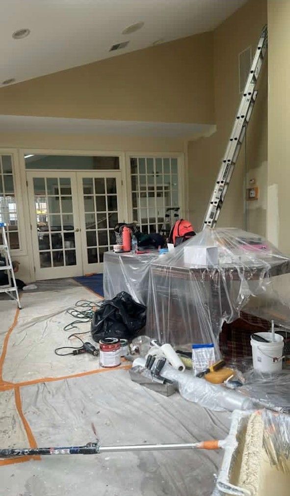 Patio under renovation with tools, ladders, paint buckets, and plastic-covered furniture near French doors