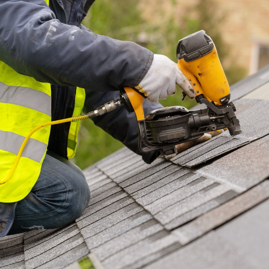 Worker using a yellow roofing nail gun on gray shingles