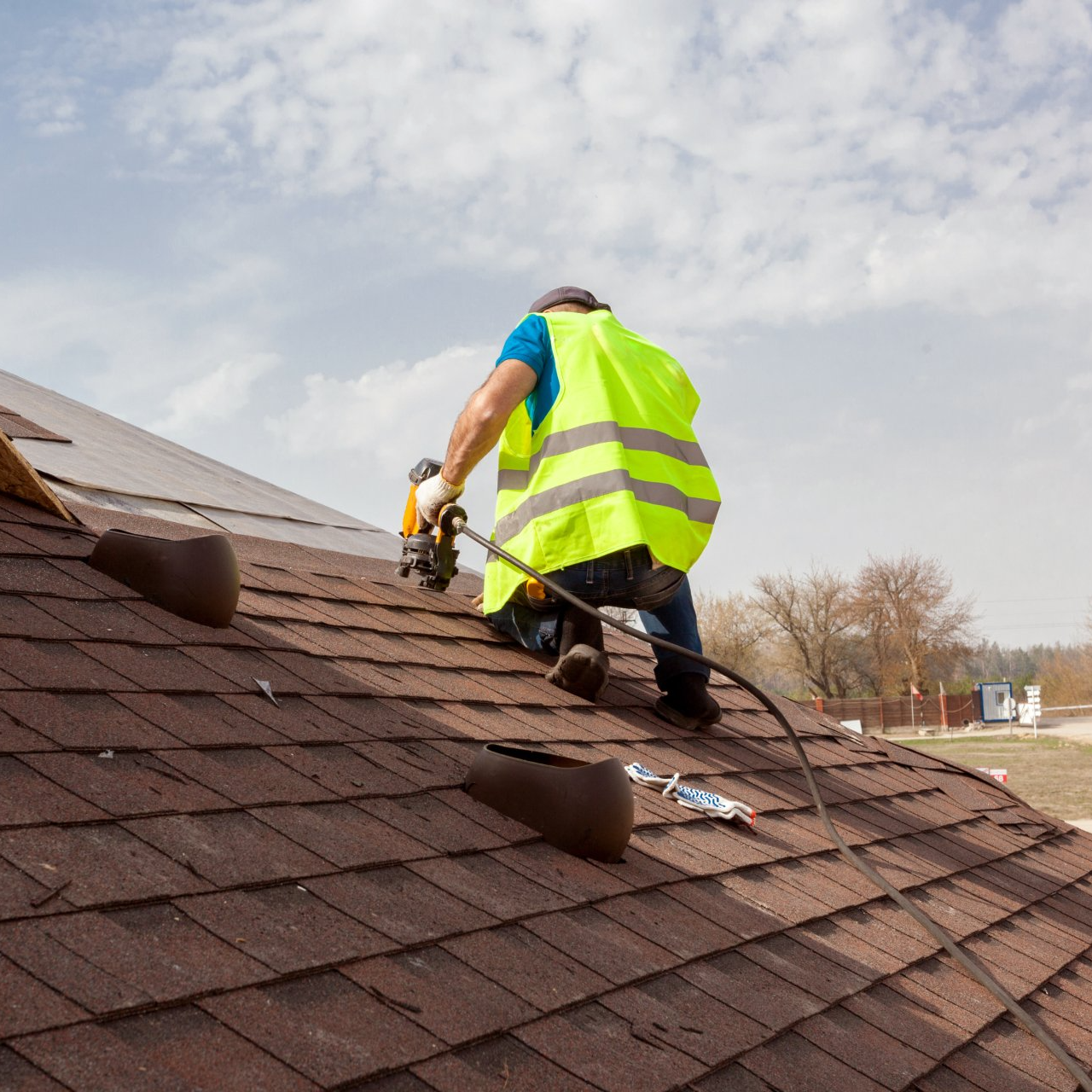 Worker in a neon safety vest repairs a shingled roof with a power drill.