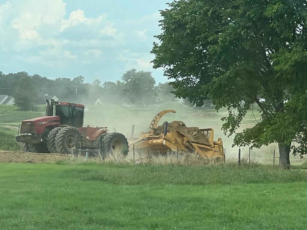 A red tractor pulls a large, yellow earth-moving scraper across a dusty, grassy field.