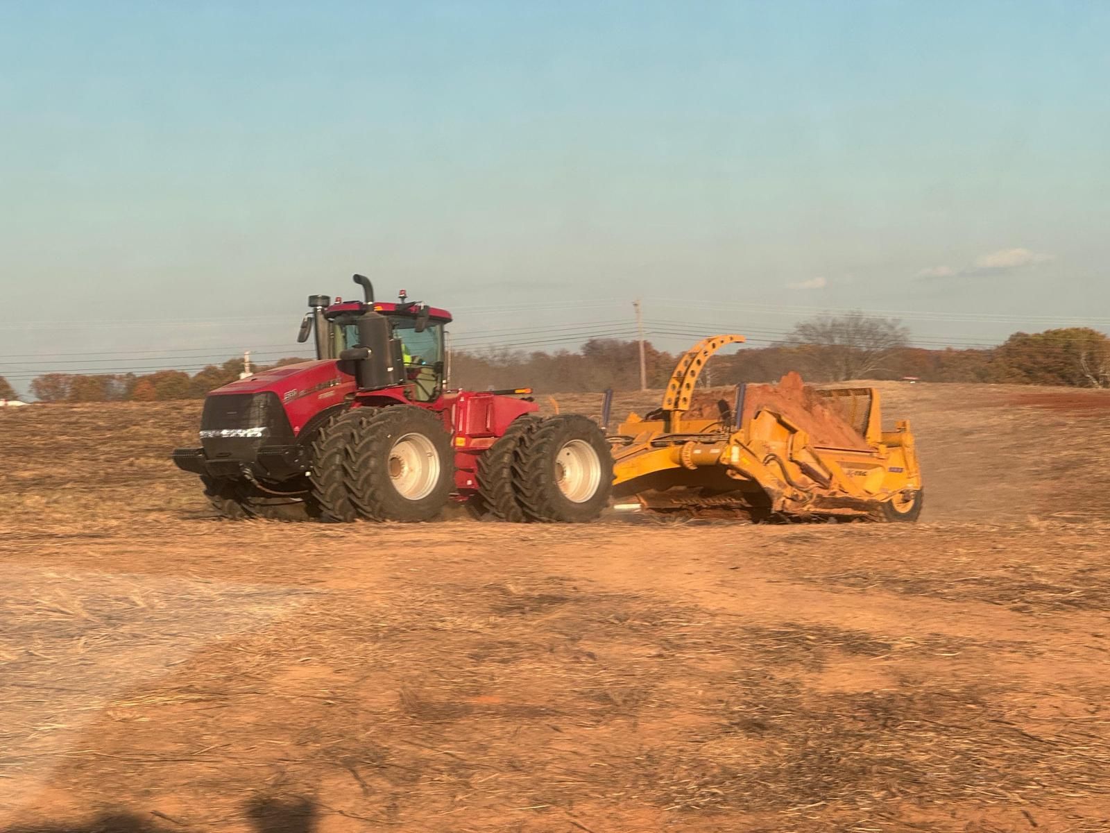 A red Case IH tractor pulling a large yellow pull-behind earth scraper across a dry, brown field.