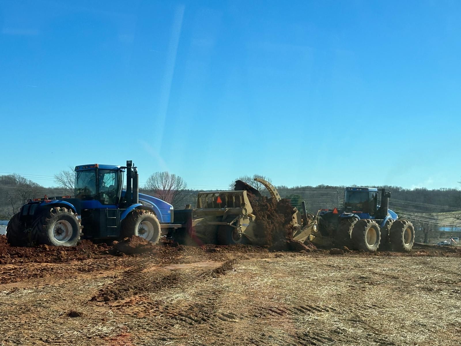 Two blue tractors operate heavy earth-moving equipment on a dirt construction site under a clear blue sky.