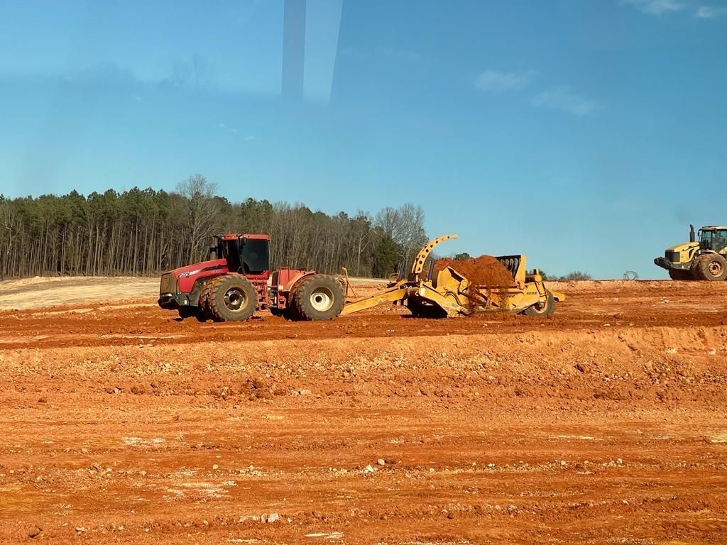 A red tractor pulls a yellow scraper filled with dirt across a large, orange-soiled construction site under a blue sky.