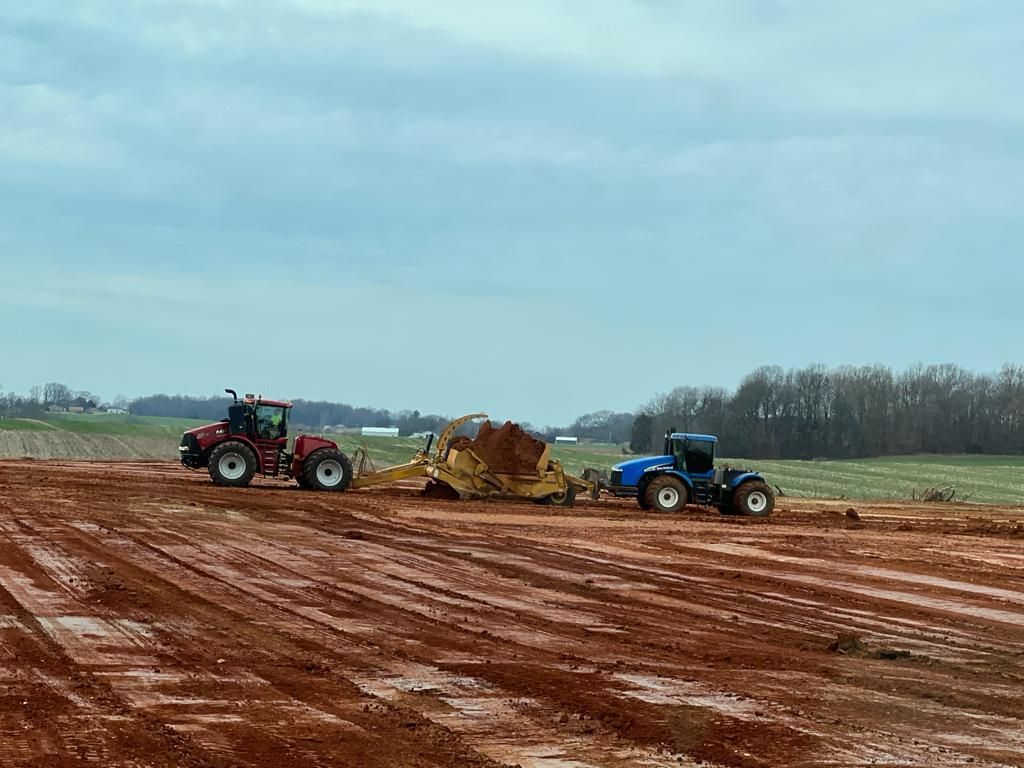 Two tractors pull a large yellow earthmoving scraper across a field of red soil under a cloudy sky.