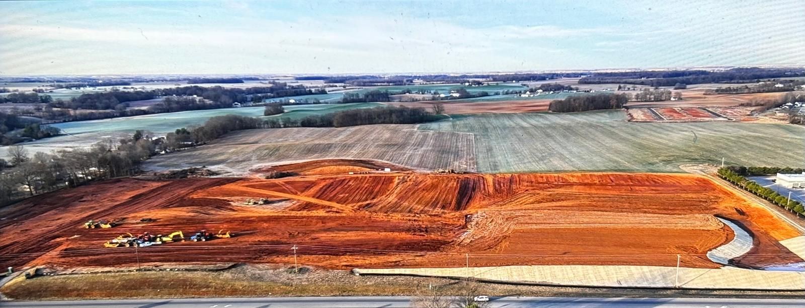 An aerial view of a construction site featuring a large, cleared plot of red dirt surrounded by fields and trees.