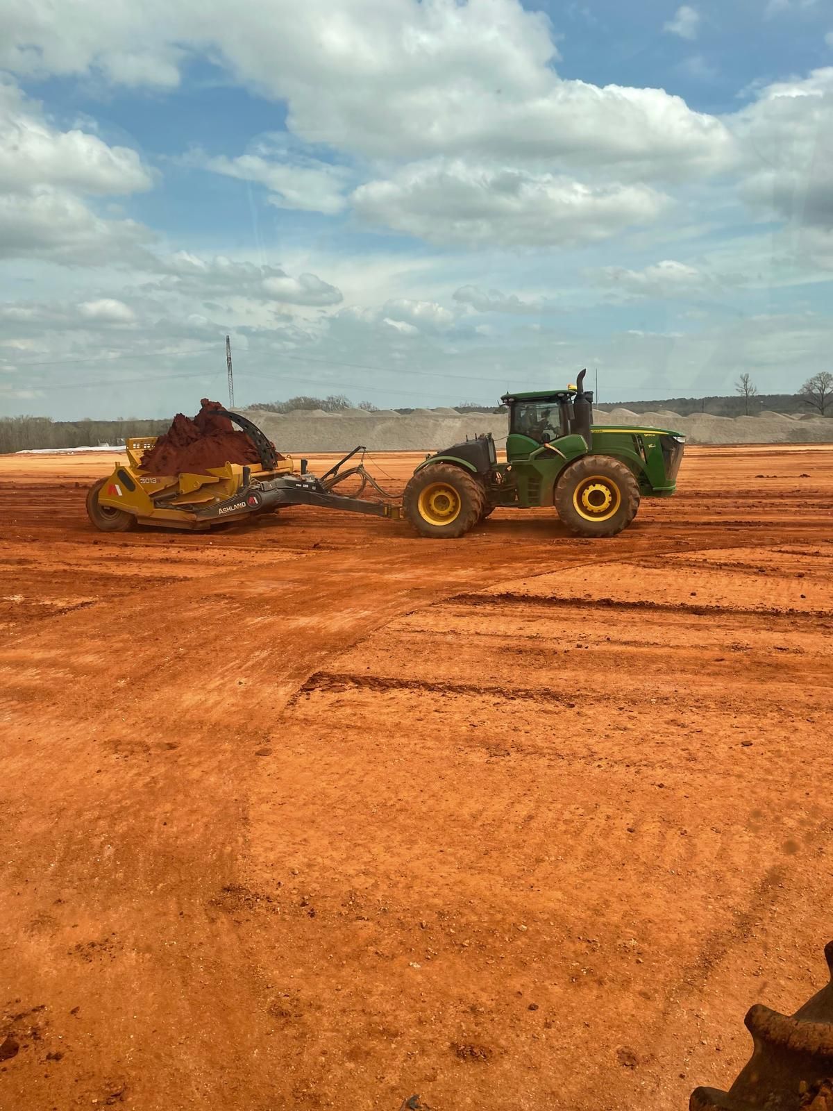 A green John Deere tractor pulls a large yellow earth scraper across a vast, flat red-dirt construction site.