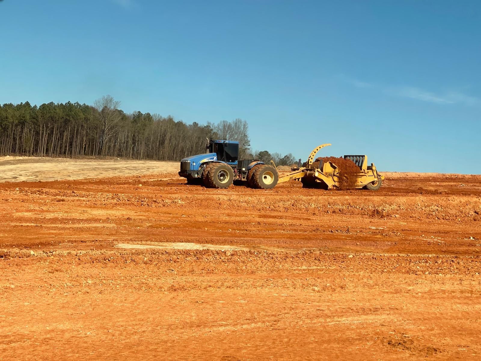 A blue tractor pulls a yellow scraper across a flat, red dirt construction site under a clear blue sky.