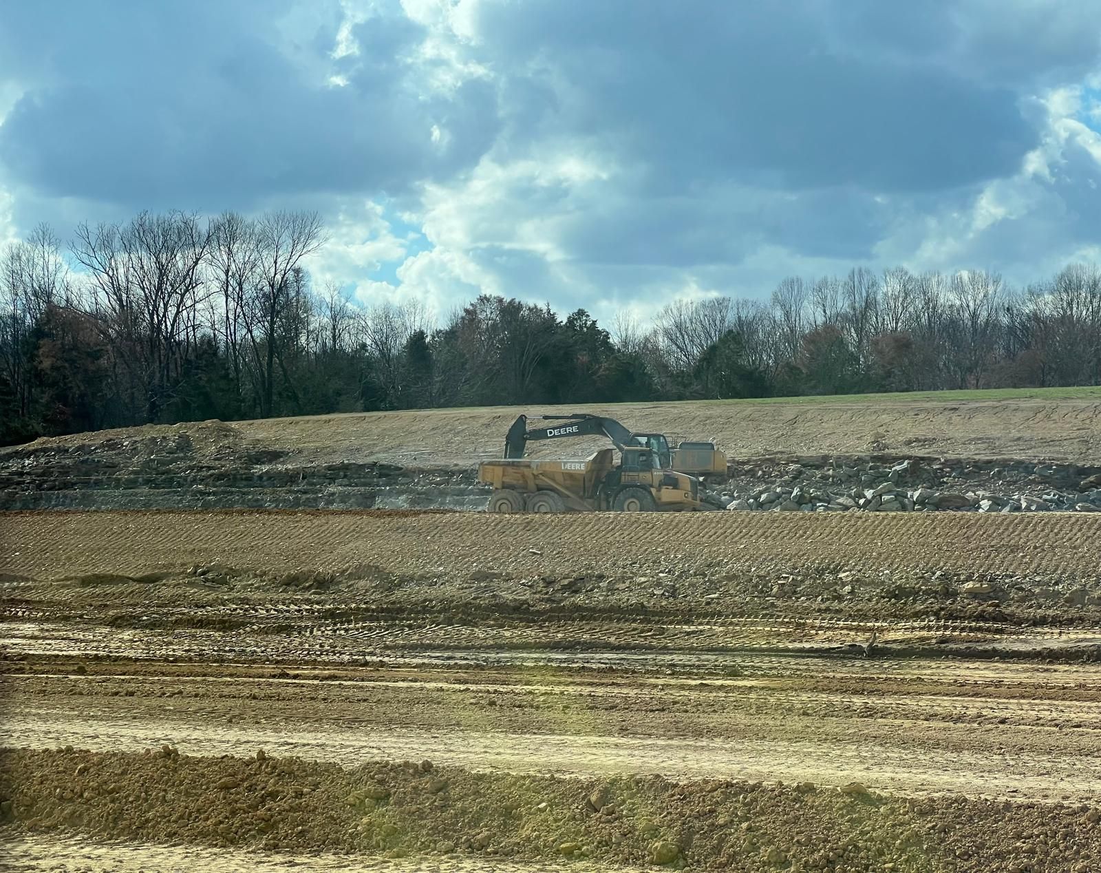 A yellow earth-moving machine works on a muddy, cleared field under a partly cloudy sky.