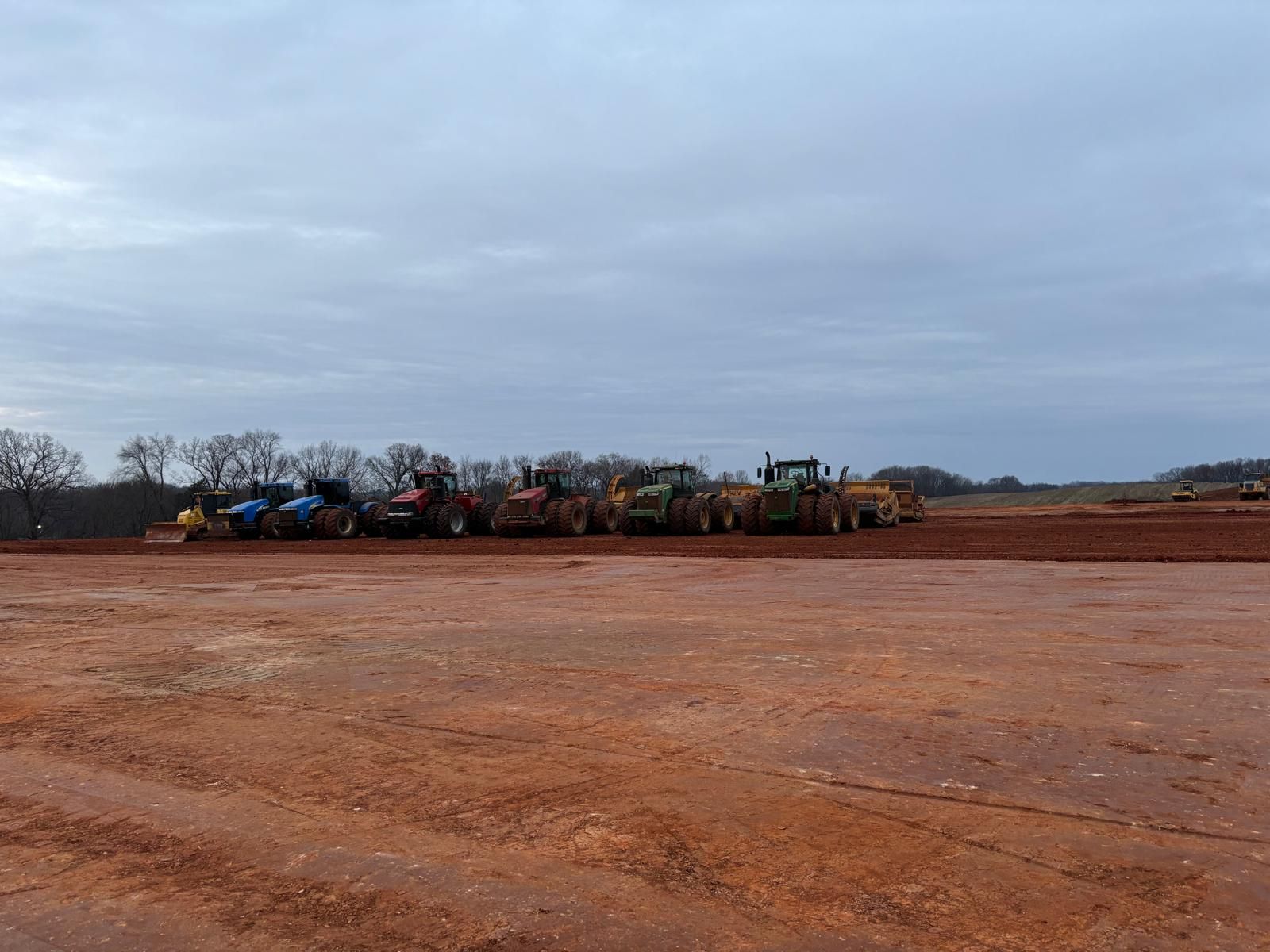 A line of various tractors and heavy machinery parked on a large, flat, red-dirt construction site under a cloudy sky.