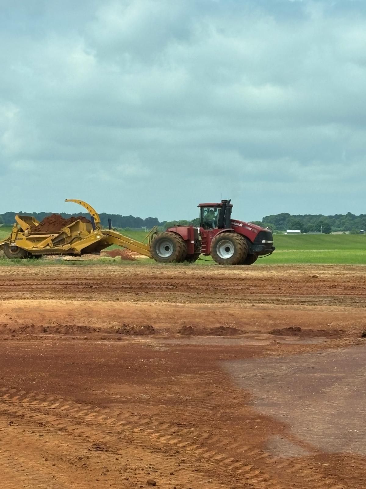A red tractor pulls a yellow dirt scraper across a bare, reddish-brown construction field under a cloudy sky.