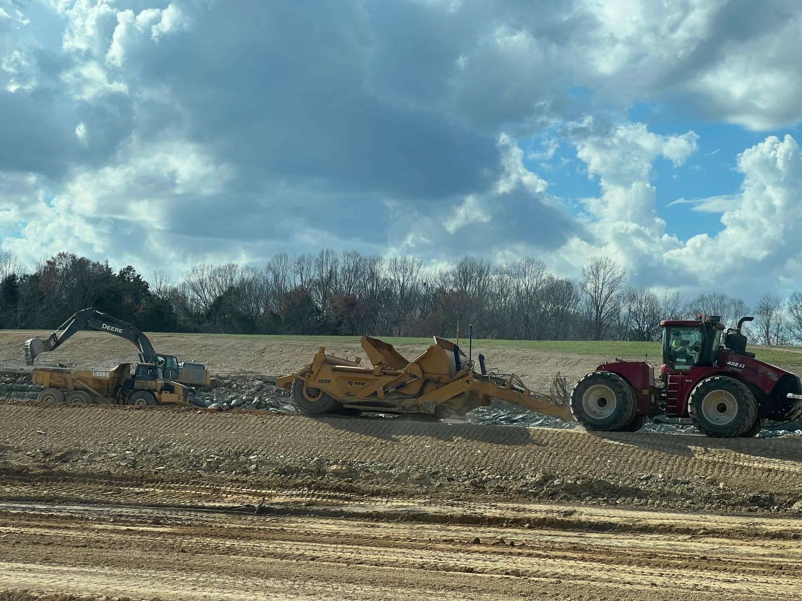 An excavator and a tractor pulling a scraper work on a muddy construction site under a cloudy blue sky.