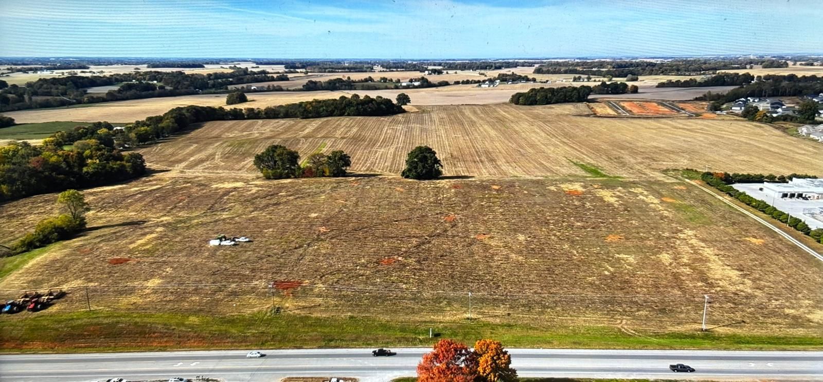 An aerial view of a large, dry, harvested field bordering a road with some trees and distant commercial buildings.