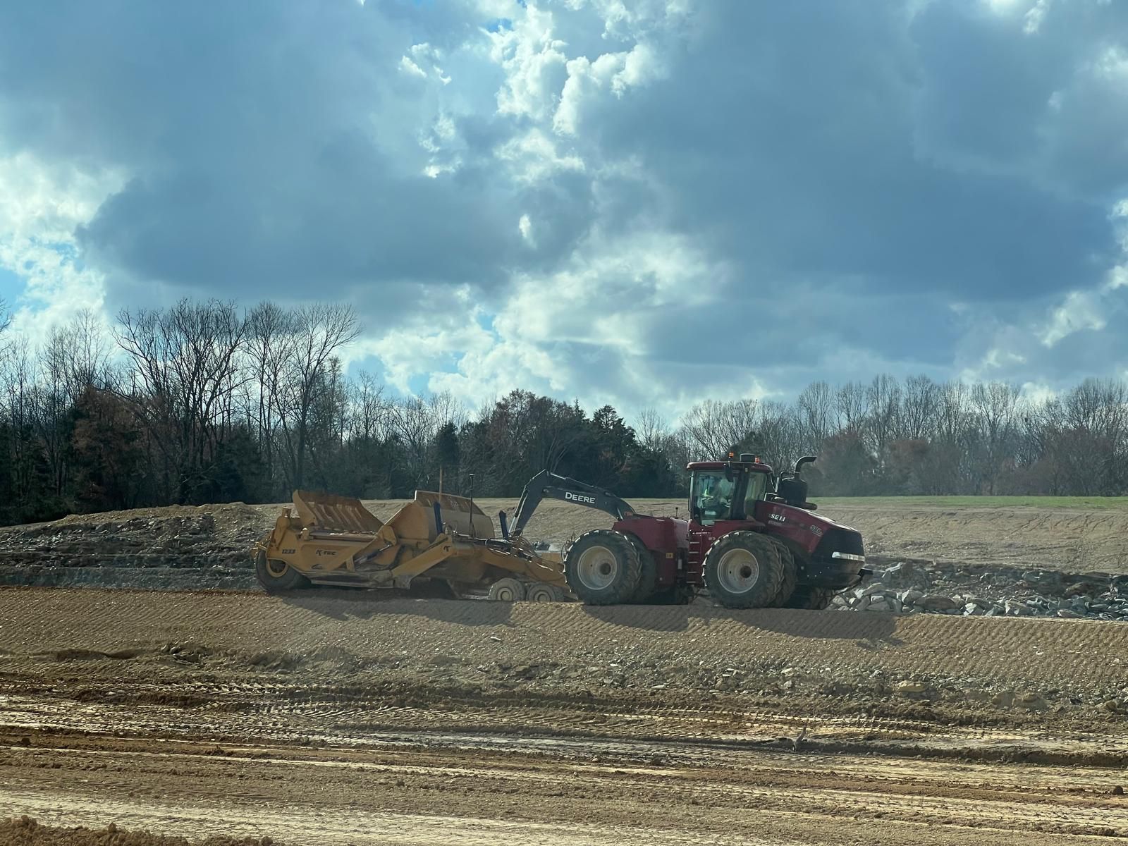 A red tractor pulls a yellow scraper through a dirt construction site under a cloudy sky.