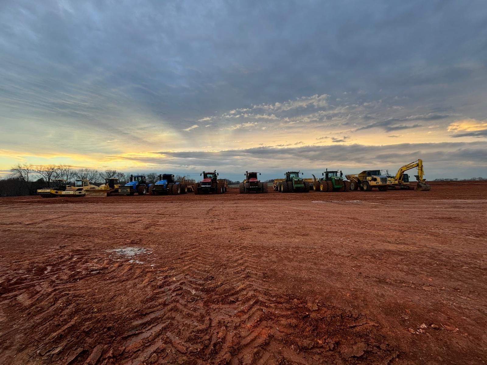 A line of heavy construction machinery parked on a reddish dirt field under a cloudy sunset sky.