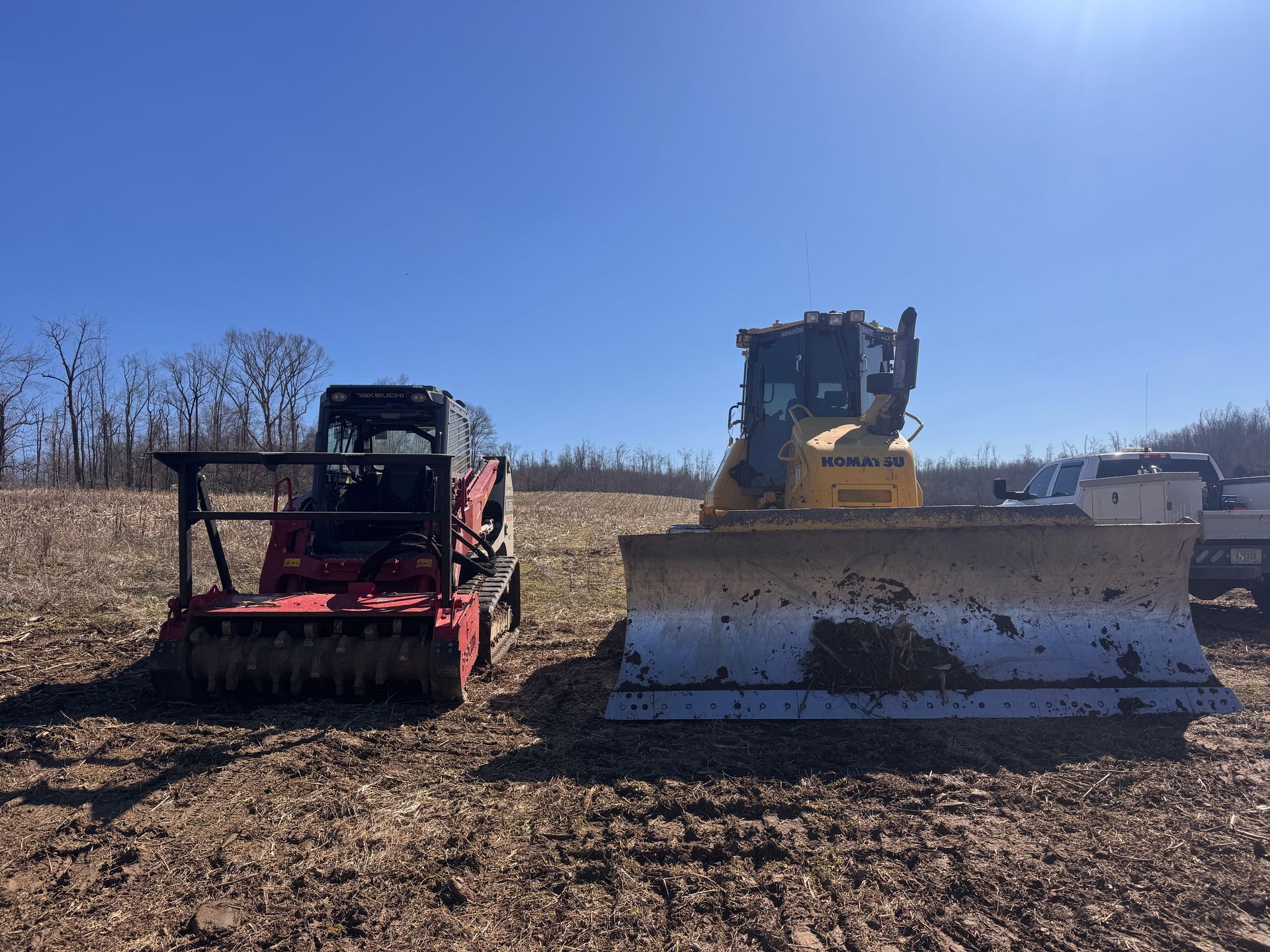 A red skid steer with a forestry mulcher and a yellow bulldozer parked side-by-side on cleared, open land under a blue sky.
