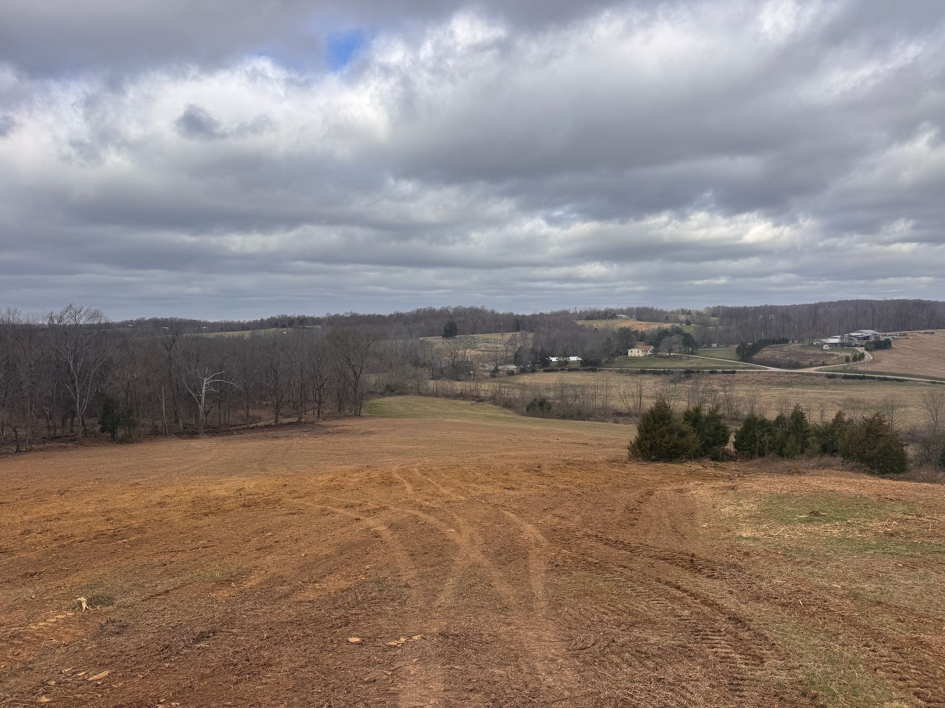 A vast, sloping field of bare brown earth leading toward a distant treeline and farmhouse under a cloudy, overcast sky.