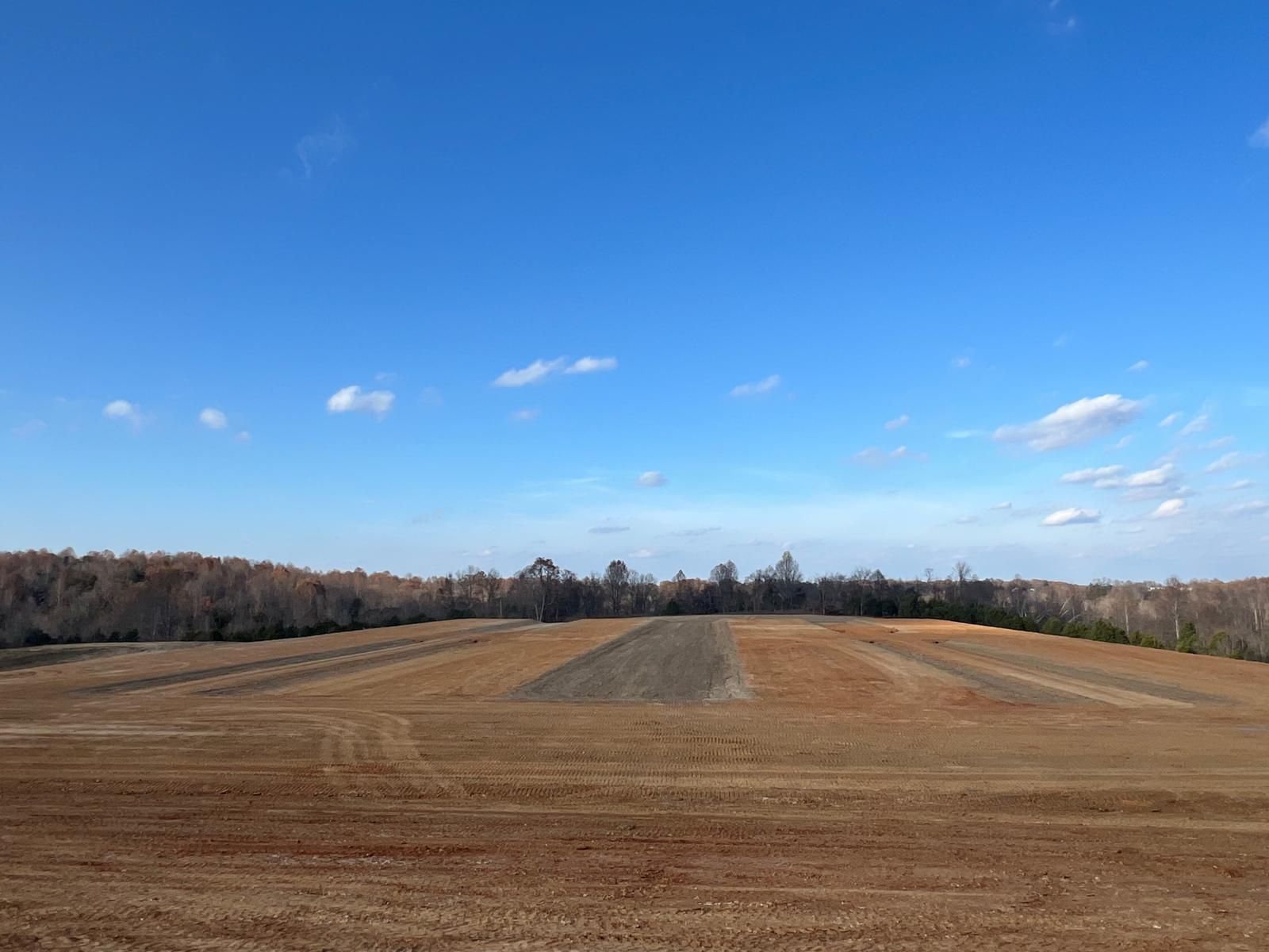 A wide, freshly plowed farm field with rows of brown soil under a bright blue sky with light clouds and a treeline.