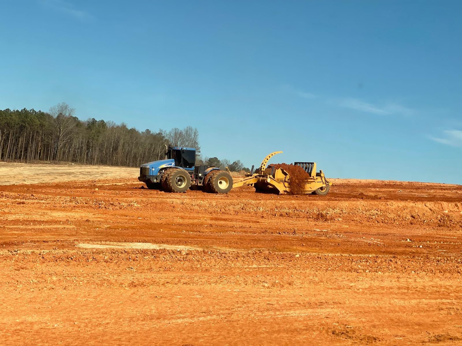 A blue tractor pulls a yellow earthmover across a barren, red-dirt construction site under a clear blue sky.