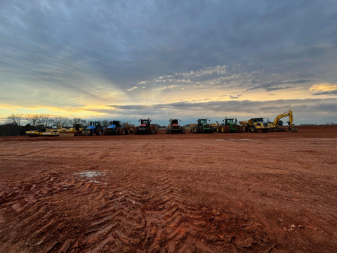 A line of heavy construction machinery parked on a reddish dirt field under a cloudy sunset sky.