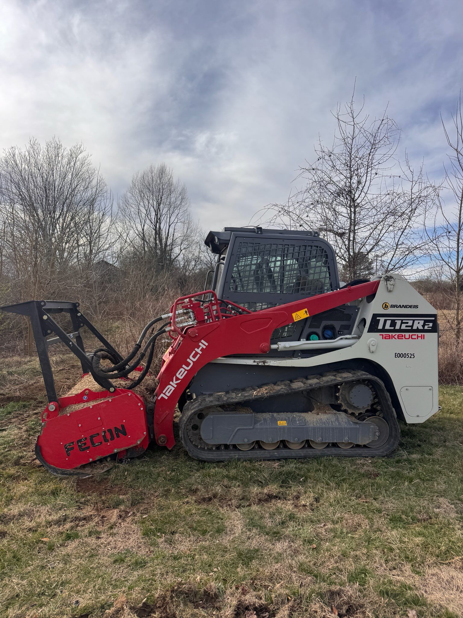 A white and red Takeuchi track loader with a forestry mulcher attachment parked in a field under a cloudy sky.