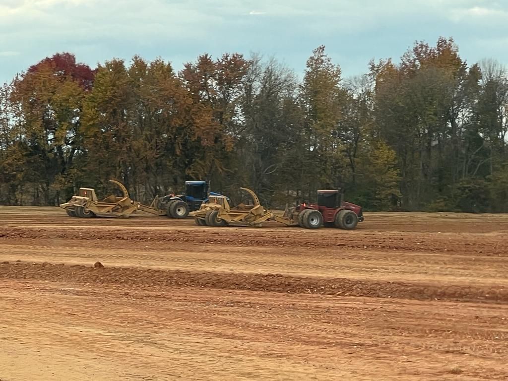 Heavy machinery, including tractors and scrapers, levels a large dirt construction site against a backdrop of trees.