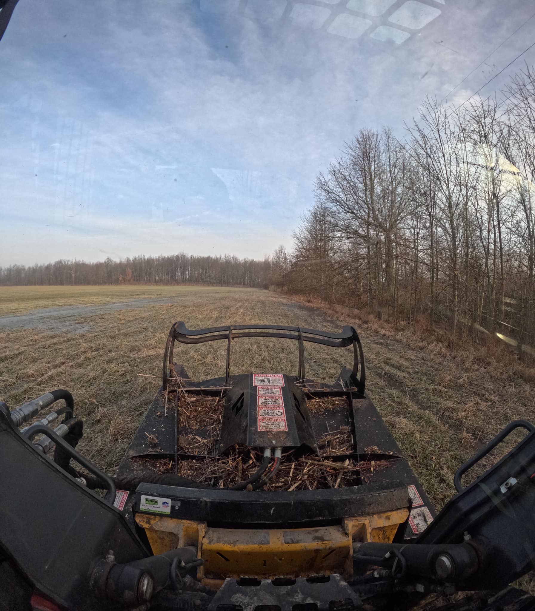 View from the cab of a yellow tractor clearing a field. Trees line the right, cloudy sky overhead.