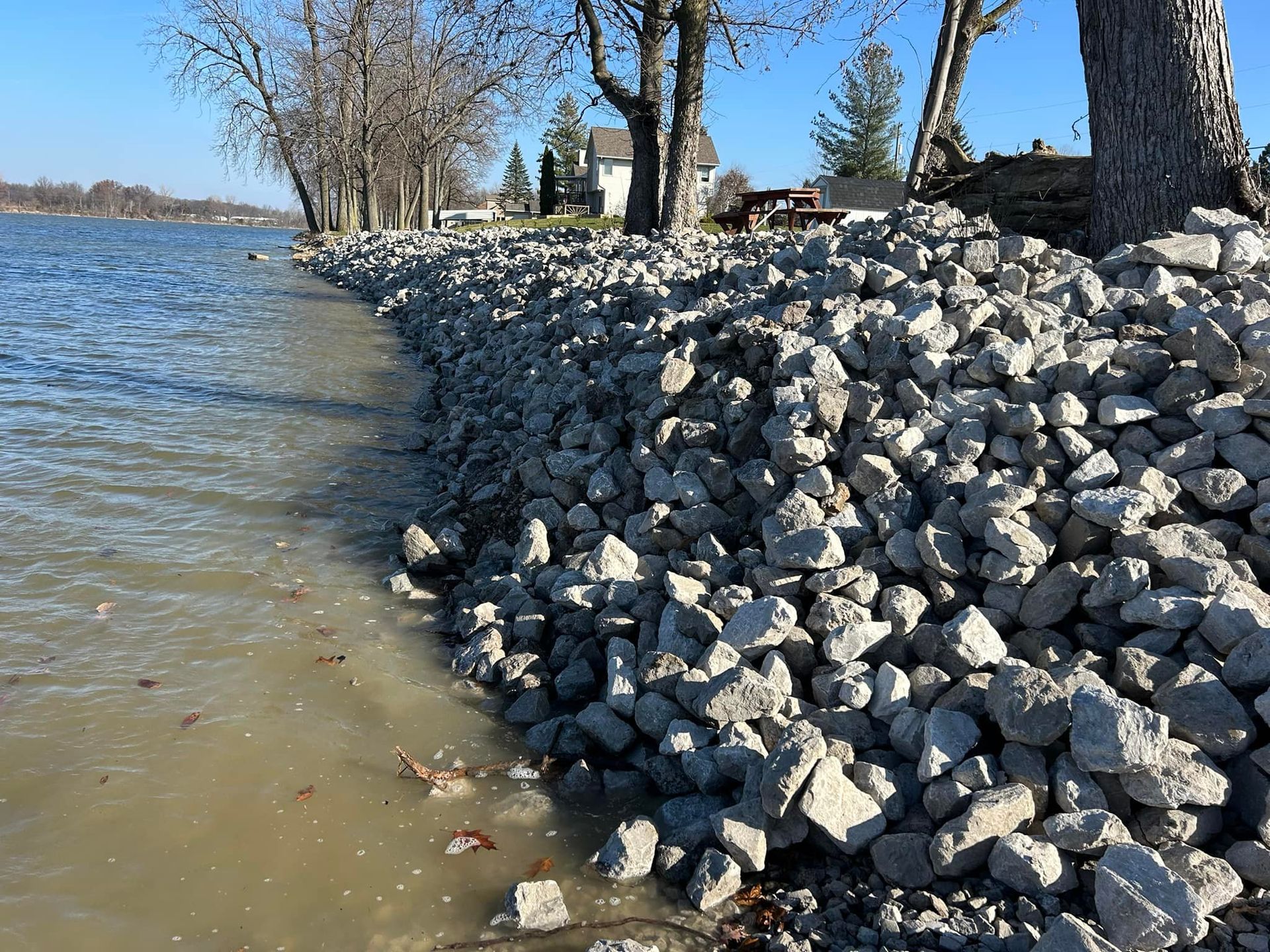 Riprap rocks protect a shoreline with bare trees and houses in the background.