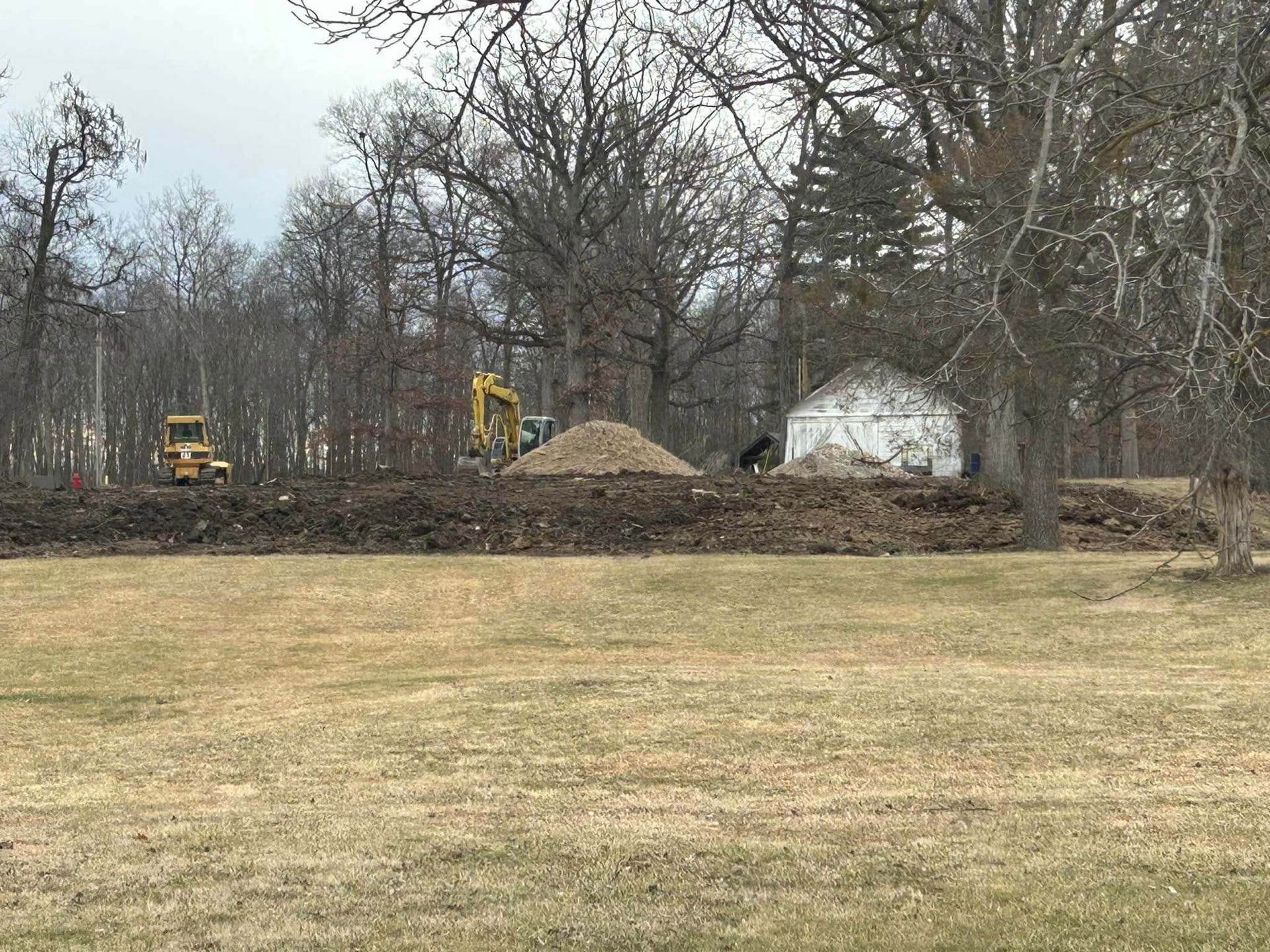 Construction site: pile of dirt with excavator and front-end loader in a grassy area, small building in background.