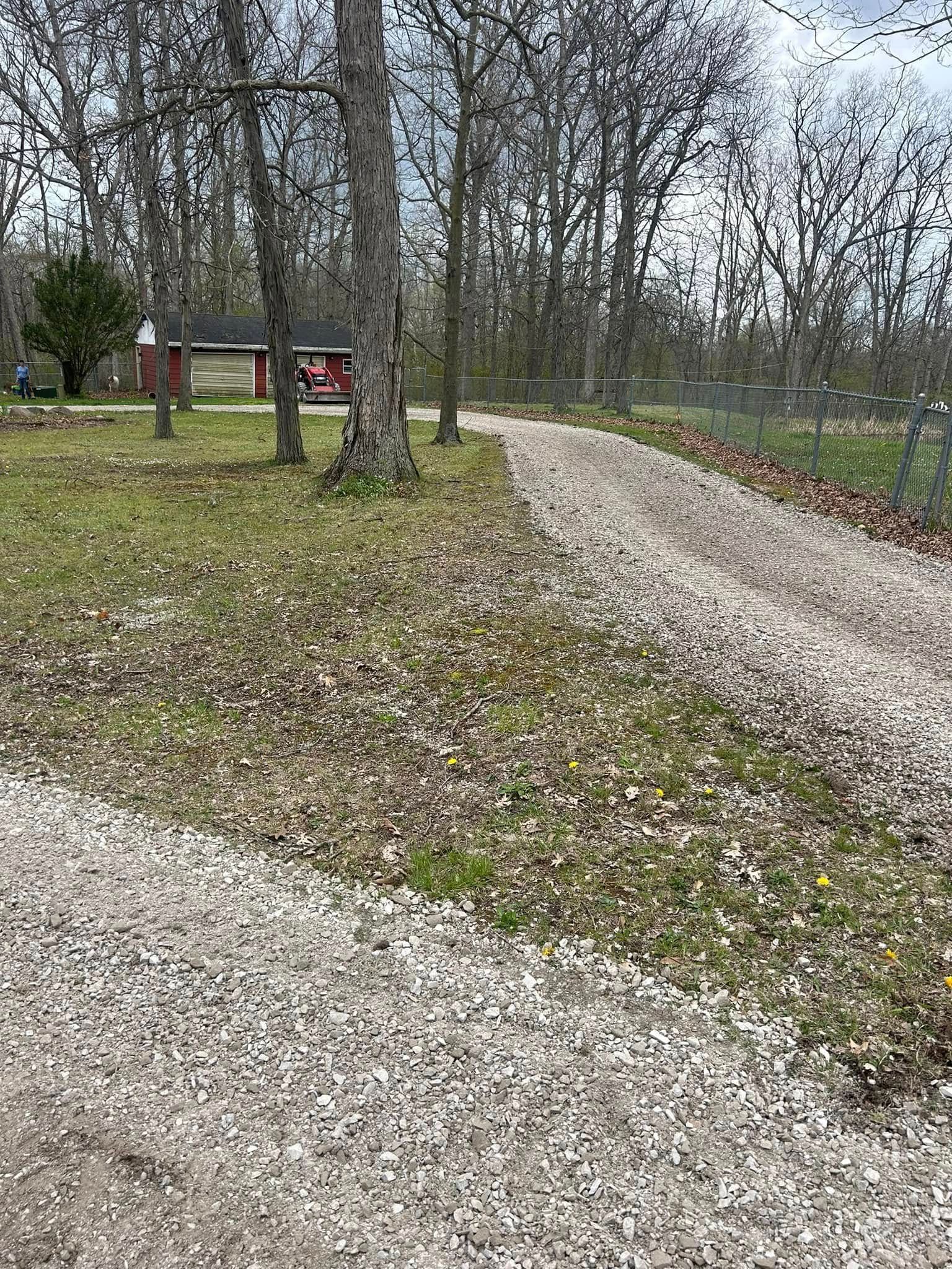 Gravel driveway leading toward a red building in a wooded area. Trees line the path, grass present.