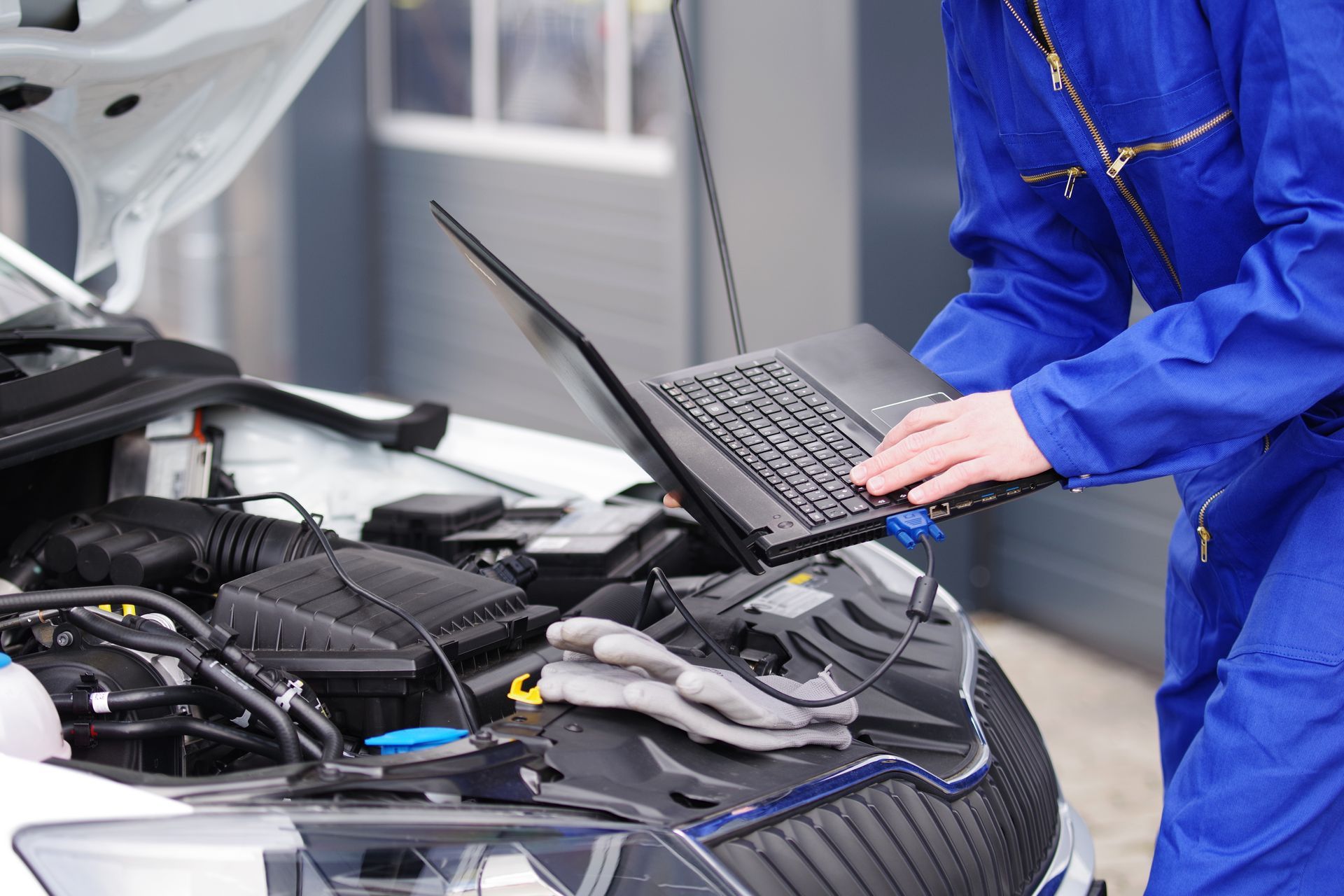 Mechanic in blue jumpsuit using a laptop to diagnose a car engine.