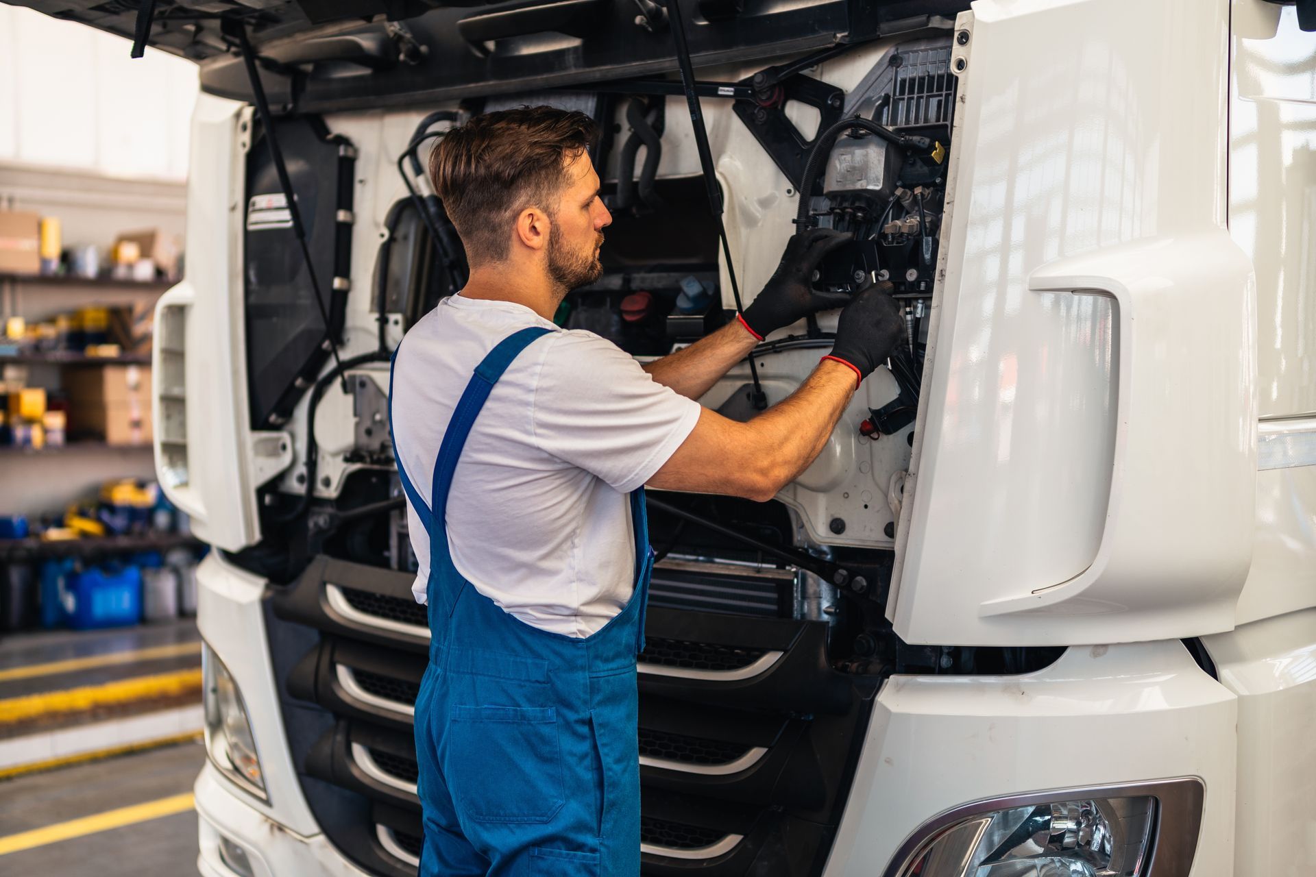Mechanic in blue overalls, fixing a truck engine with the hood open. Garage setting.