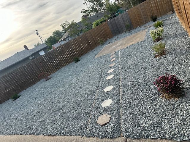 A backyard with gray gravel, a wooden fence, a concrete patio area, and a stone path leading to a small square patio.