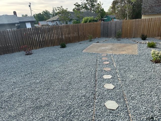 A backyard with grey gravel, a wooden fence, a concrete patio area, and a stone stepping stone path leading to it.