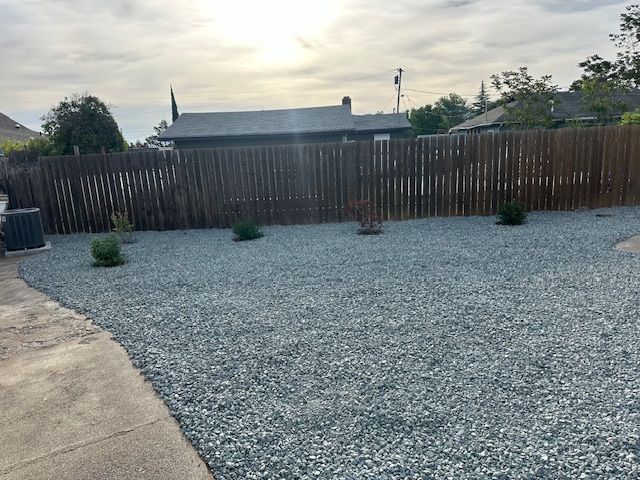 A backyard with gray gravel ground cover, a few small shrubs, and a tall wood fence against a bright sky.