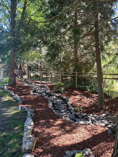 A rock-lined garden path filled with wood mulch meanders through a wooded area under a wooden fence.