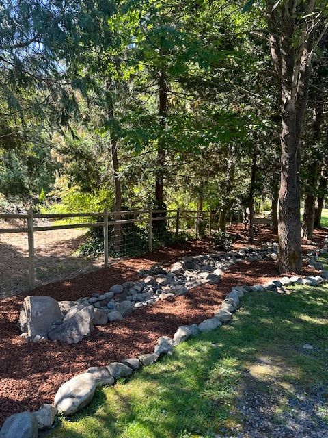 A mulched landscape area features a stone-lined dry creek bed winding through a wooded yard with a wooden fence.