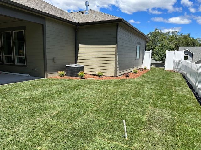A side view of a house with tan siding, a small landscaping bed with young shrubs, and a white privacy fence.