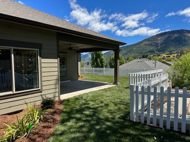 A tan house with a covered patio overlooks a grassy backyard with a white picket fence and distant mountains.