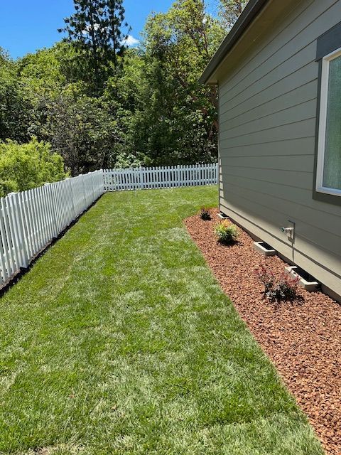 A side yard with a manicured green lawn, a red mulch garden bed with small plants, and a white picket fence.