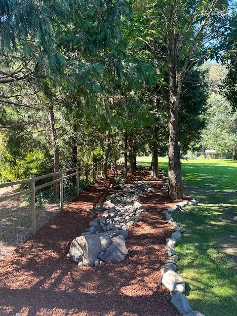 A gravel and rock dry creek bed bordered by a wooden fence, mulch, and trees, leading toward a sunny green lawn.