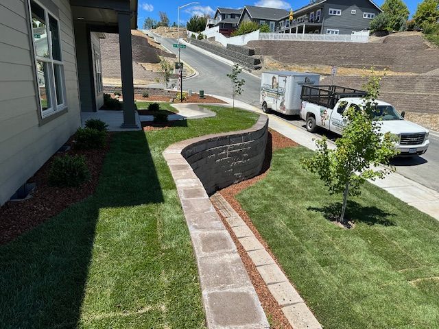 A view of a newly landscaped suburban yard with a curved stone retaining wall, fresh grass, and a truck parked on the street.