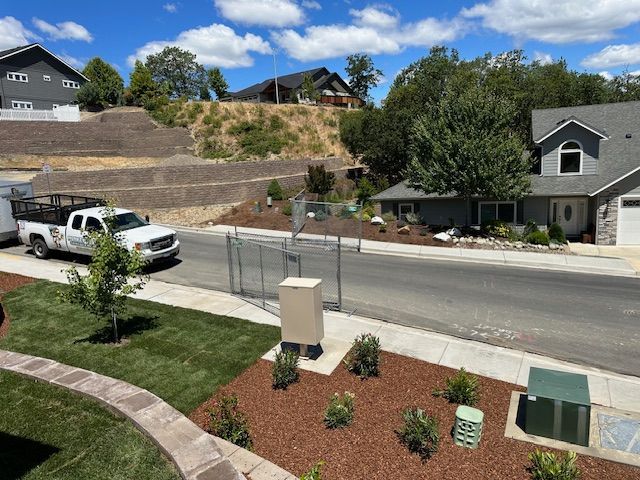A white work truck parked on a suburban street near a landscaped yard with mulch, young plants, and a tiered hill.