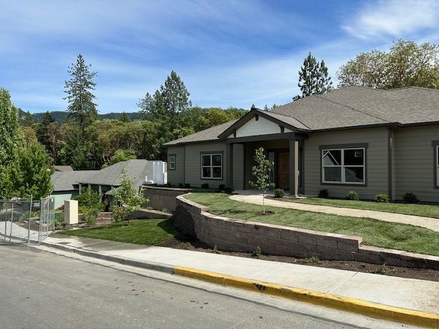 A taupe single-story building with a front porch, stone retaining wall, and green landscaping under a bright blue sky.