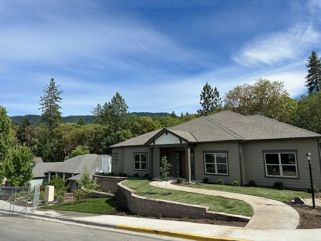 A tan, single-story house with a grey roof sits on a sloped lot with a curved concrete path and a stone retaining wall.